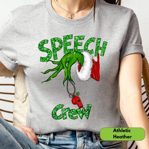 May include: A heather gray short-sleeve t-shirt featuring the green glitter text SPEECH CREW. Between the words is a graphic of a green furry hand emerging from a red and white Santa sleeve, holding a red whistle on a black lanyard.