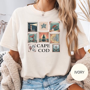 Op de afbeelding: Ivoor kleurig T-shirt met een Cape Cod ontwerp. Het ontwerp bevat negen postzegel-achtige afbeeldingen met nautische thema's, zoals een vuurtoren, zeester en zeilboot. De woorden "CAPE COD" zijn vetgedrukt.