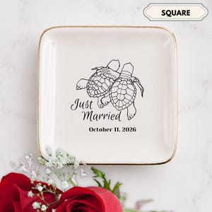 May include: A square white ceramic trinket dish with a gold-toned rim. The center features a black line drawing of two sea turtles, the text "Just Married," and the date "October 11, 2026" printed in a clear, dark font.