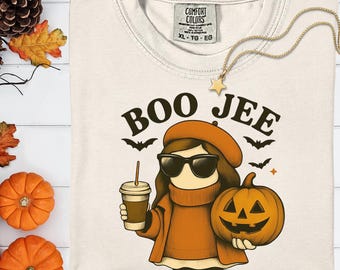 May include: Cream-colored t-shirt with a graphic of a person wearing sunglasses, an orange beret, and holding a coffee and a carved pumpkin. The text "BOO JEE" is above the graphic, with bats and stars. A gold star necklace is draped on the shirt.