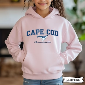 May include: A light pink hooded sweatshirt featuring a front kangaroo pocket. The chest displays the text CAPE COD in dark blue block letters, a graphic of a blue whale, and the word Massachusetts written in a cursive script font below the whale.