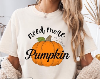 Puede incluir: Una camiseta de manga corta color marfil con un gráfico de una calabaza naranja con una hoja verde y un tallo marrón. El texto "need more" está impreso en negro sobre la calabaza y la palabra "Pumpkin" está impresa en negro debajo.
