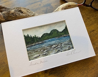 May include: A watercolor painting of a landscape scene with a river, rocks, and trees. The painting is framed in a white mat and has the text "Acadia, Maine" written in cursive at the bottom. The artist's signature is in the bottom right corner.