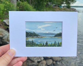 May include: A watercolor painting of a coastal scene with a body of water, trees, and birds flying overhead. The painting is framed in a white mat.
