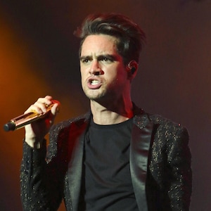 May include: A man with short brown hair and a black blazer with a sparkly texture is singing into a gold microphone. He is wearing a black t-shirt underneath the blazer.