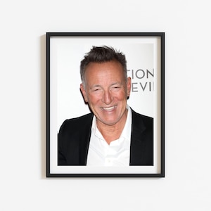 May include: A black and white photo of a man with short brown hair and a black suit jacket. He is smiling and looking at the camera. The background is white.