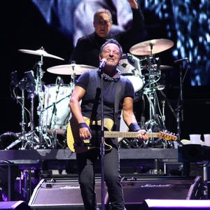 May include: A musician playing a yellow electric guitar on stage with a microphone and a drummer behind him.