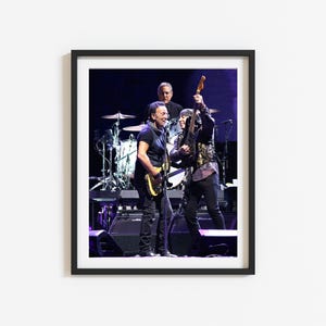 May include: A black and white photo of three musicians on stage performing. The musicians are playing guitars and singing. The stage is lit with purple and blue lights.