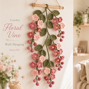 Op de afbeelding: Een gehaakte bloemenrank wandhanger met roze en donkerroze bloemen en groene bladeren, opgehangen aan een houten stok. De tekst luidt Crochet Floral Vine Wall Hanging Pattern PDF. Het ontwerp bestaat uit meerdere stengels en bloesems.