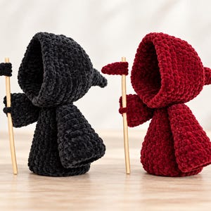 May include: Two crocheted figures made of soft, plush yarn, one in black and one in deep red. Each figure wears a hooded cloak and holds a small, light-colored wooden staff topped with a matching crocheted shape resembling a scythe blade.