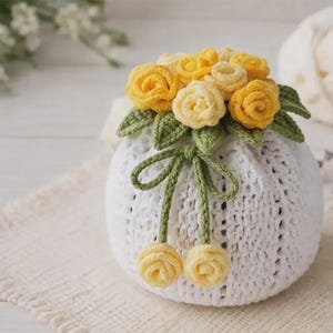 May include: A white crocheted pouch adorned with yellow and cream-colored crocheted roses and green leaves. The pouch has a drawstring closure with two yellow rosebud accents. The handmade item is displayed on a white surface.