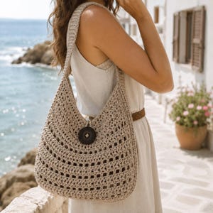Può includere: Una borsa hobo all'uncinetto color beige con un motivo a maglia aperta e una chiusura con un singolo bottone circolare marrone scuro al centro. La borsa presenta un'ampia tracolla ed è realizzata in filato testurizzato con un punto regolare.