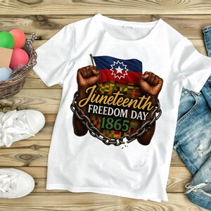 May include: White t-shirt featuring a graphic design for Juneteenth Freedom Day. The design includes a flag with a star, fists, chains, and the text "Juneteenth Freedom Day 1865". The shirt is displayed on a wooden surface.