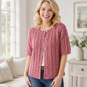 May include: A dusty rose short-sleeved cardigan featuring a textured cable knit pattern, a round neckline, and a front button closure with matching circular buttons. The garment is worn over a white top and blue denim jeans, showcasing a knit texture.
