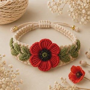 May include: A beige macrame bracelet featuring a central red poppy flower with a dark green and black center, flanked by embroidered green leaves on each side. The bracelet has an adjustable sliding knot closure with small gold-toned beads.