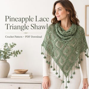 Könnte beinhalten: Ein salbeigrünes Dreieckstuch mit einem Ananas-Spitzen-Häkelmuster und baumelnden Bommel-Fransen an den Rändern. Der Text Pineapple Lace Triangle Shawl Crochet Pattern PDF Download ist auf der linken Seite des Bildes zu sehen.