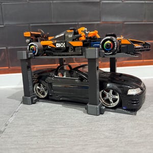 May include: A black convertible car model is displayed on a gray stand, with a black and orange race car model on top. The race car has the word "chrome" printed on it.