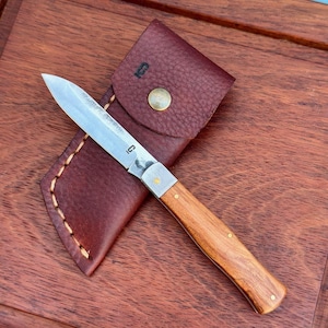 May include: A folding pocket knife with a polished steel blade featuring a small logo, a light brown wooden handle with brass pins, and a dark burgundy textured leather sheath with a brass snap button and a small stamped logo on the flap.