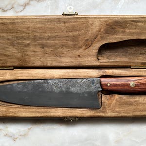 Könnte beinhalten: Ein großes Kochmesser mit dunkler Klinge und Holzgriff liegt in einer offenen Holzkiste. Das Messer hat eine strukturierte Klinge und einen rotbraunen Griff mit Messingakzent. Die Kiste ist aus hellbraunem Holz gefertigt und hat Messingscharniere.