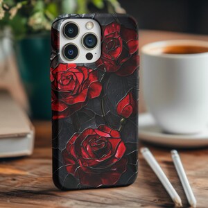 Pu&ograve; includere: Una custodia per iPhone nera con un design di rose rosse su uno sfondo scuro. La custodia ha un ritaglio per l'obiettivo della fotocamera e un bordo rialzato attorno allo schermo.