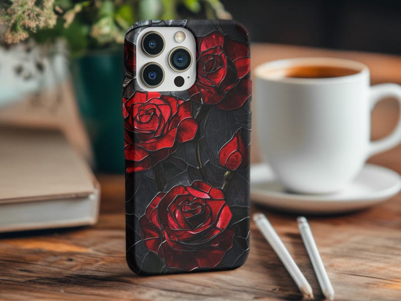 Pu&ograve; includere: Una custodia per iPhone nera con un design di rose rosse su uno sfondo scuro. La custodia ha un ritaglio per l'obiettivo della fotocamera e un bordo rialzato attorno allo schermo.