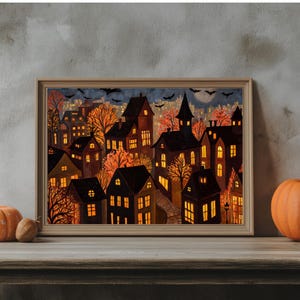 May include: A framed print depicting a nighttime cityscape with illuminated houses, trees, and bats. The artwork features warm orange and yellow hues, evoking a cozy autumn or Halloween theme. Two pumpkins sit in front of the frame.