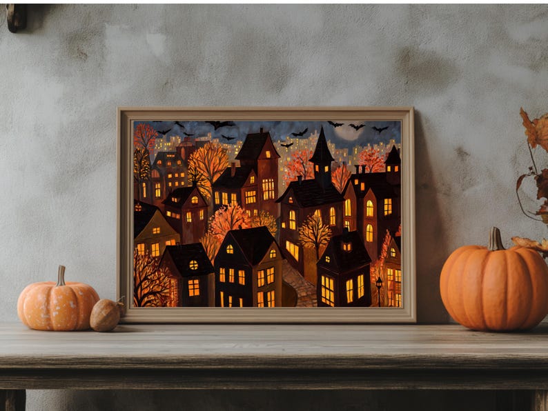 May include: A framed print depicting a nighttime cityscape with illuminated houses, trees, and bats. The artwork features warm orange and yellow hues, evoking a cozy autumn or Halloween theme. Two pumpkins sit in front of the frame.