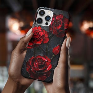 Pu&ograve; includere: Una custodia per telefono nera con un design di rose rosse su uno sfondo scuro. La custodia ha un ritaglio per l'obiettivo della fotocamera e un bordo nero.