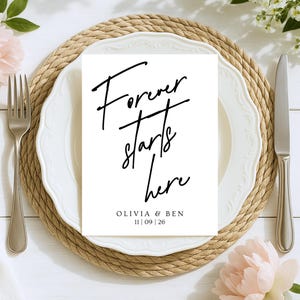May include: A white card with the text "Forever starts here" in black script, placed on a white plate with a decorative edge. The plate sits on a woven placemat, with a fork and knife on either side. The names Olivia & Ben and the date 11|09|26 are below the text.
