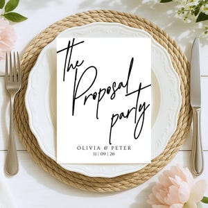 May include: A white card with black script that reads "The Proposal Party" sits on a white plate with a decorative edge. The card also includes the names "Olivia & Peter" and the date "11 | 09 | 26". The plate rests on a woven placemat.