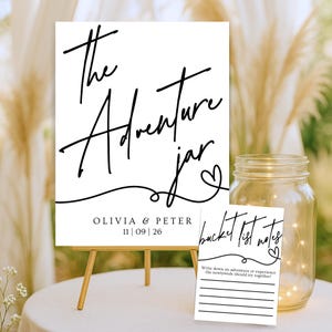May include: A white sign on a small wooden easel with the words "The Adventure Jar" in black script. Below the text is the names "Olivia & Peter" and the date "11|09|26". A clear glass jar and a small card with "bucket list notes" are also visible.