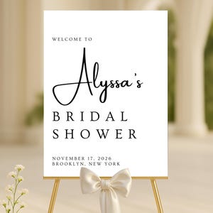 May include: A white sign on a gold easel, announcing "Welcome to Alyssa's Bridal Shower." The text is in black script and block letters. The date is November 17, 2026, in Brooklyn, New York. A white bow adorns the easel.