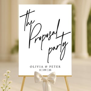May include: A white sign with the words "The Proposal Party" in elegant black script. Below, it reads "OLIVIA & PETER" and the date "11|09|26". The sign is displayed on a gold easel, adorned with a cream-colored bow.