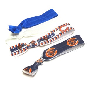 May include: Three hair ties with a Chicago Bears theme. One is white with orange and blue zigzags, one is blue, and one is navy blue with orange and white bears.