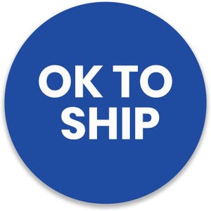 May include: A round, blue sticker with the white text "OK TO SHIP". The sticker has a white border and a slight shadow, suggesting a raised surface. The text is in a bold, sans-serif font.