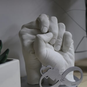 May include: A white plaster hand cast of two hands holding each other, mounted on a silver base with an infinity symbol and the words "My & Love".