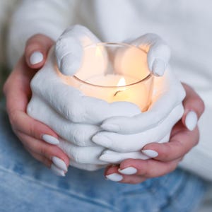 May include: White ceramic hand-shaped candle holder with a glass candle inside. The candle is lit and the flame is visible.