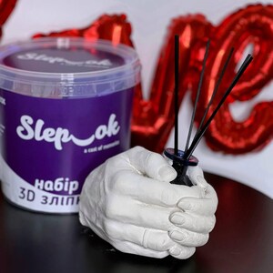 Könnte beinhalten: Eine weiße Gipshandskulptur hält eine schwarze Glasflasche mit Diffusorstäbchen. Die Flasche ist mit "Sleep ok" beschriftet und die Handskulptur ist ein 3D-Abguss. Der Hintergrund ist ein roter herzförmiger Ballon.