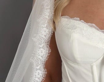 May include: A white corset top featuring a sweetheart neckline with delicate lace trim and a matching sheer veil with an intricate floral lace border. The corset includes vertical boning details and a tie-front closure at the center neckline.