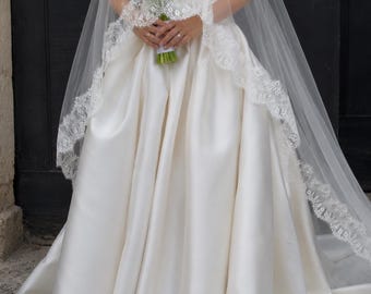 May include: A floor-length ivory satin wedding dress with a strapless bodice and a full, pleated skirt. A sheer, floor-length veil with intricate white lace trim along the edges is draped over the dress. A bouquet of white calla lilies is held.