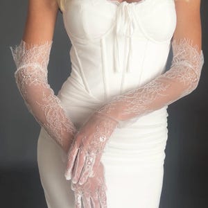 May include: A white, strapless corset-style bodice dress with a sweetheart neckline. The dress is paired with long, sheer white lace gloves. The gloves feature intricate floral lace patterns and extend to the upper arm, creating an elegant look.
