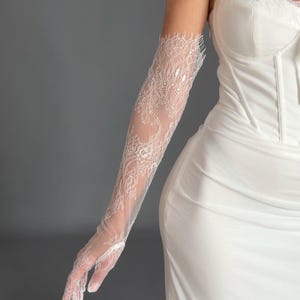 May include: Elegant white lace gloves with a floral design. The gloves are long, reaching up the arm, and are worn with a white dress. The lace is detailed, creating a sophisticated and formal appearance.