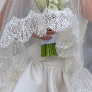 May include: A white wedding veil featuring an intricate scalloped lace border with floral patterns, draped over a bouquet of white calla lilies with green stems wrapped in white ribbon, worn with a smooth, solid white satin dress.