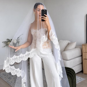 May include: A long white bridal veil featuring intricate scalloped lace trim along the edges, paired with a strapless lace corset top and wide-leg white trousers. The sheer tulle veil drapes over the head and shoulders, showcasing detailed floral embroidery patterns on the lace border.