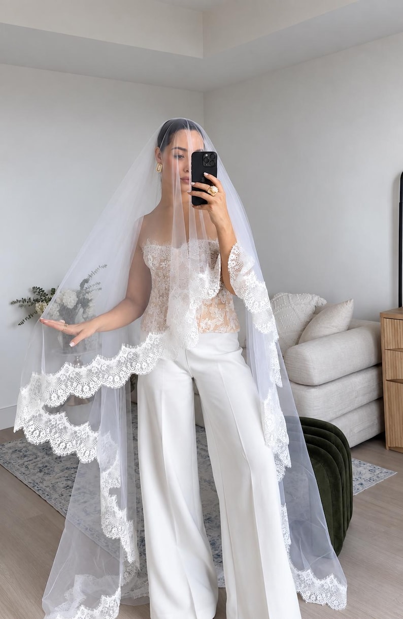 May include: A long white bridal veil featuring intricate scalloped lace trim along the edges, paired with a strapless lace corset top and wide-leg white trousers. The sheer tulle veil drapes over the head and shoulders, showcasing detailed floral embroidery patterns on the lace border.