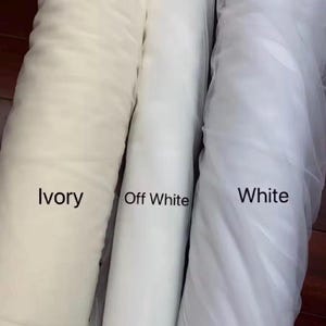May include: Three rolls of fabric in shades of white. The rolls are labelled "Ivory", "Off White", and "White". The fabric appears to be a sheer material, possibly tulle or netting, with a soft, flowing texture. The image is well-lit, highlighting the different tones.