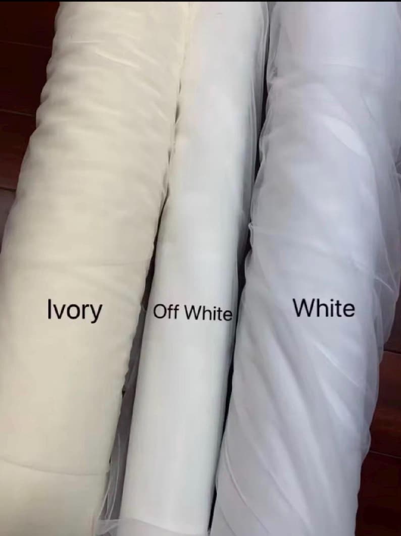 May include: Three rolls of fabric in shades of white. The rolls are labelled "Ivory", "Off White", and "White". The fabric appears to be a sheer material, possibly tulle or netting, with a soft, flowing texture. The image is well-lit, highlighting the different tones.