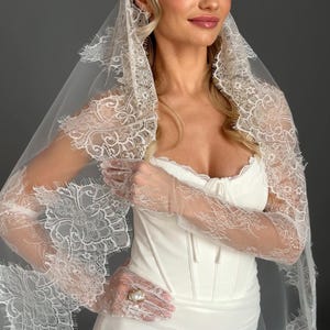 May include: A white wedding dress with a corset bodice and a fitted skirt. The bride is wearing a lace veil and lace gloves. The gloves have a pearl ring on the ring finger. The veil and gloves have a delicate floral lace design.