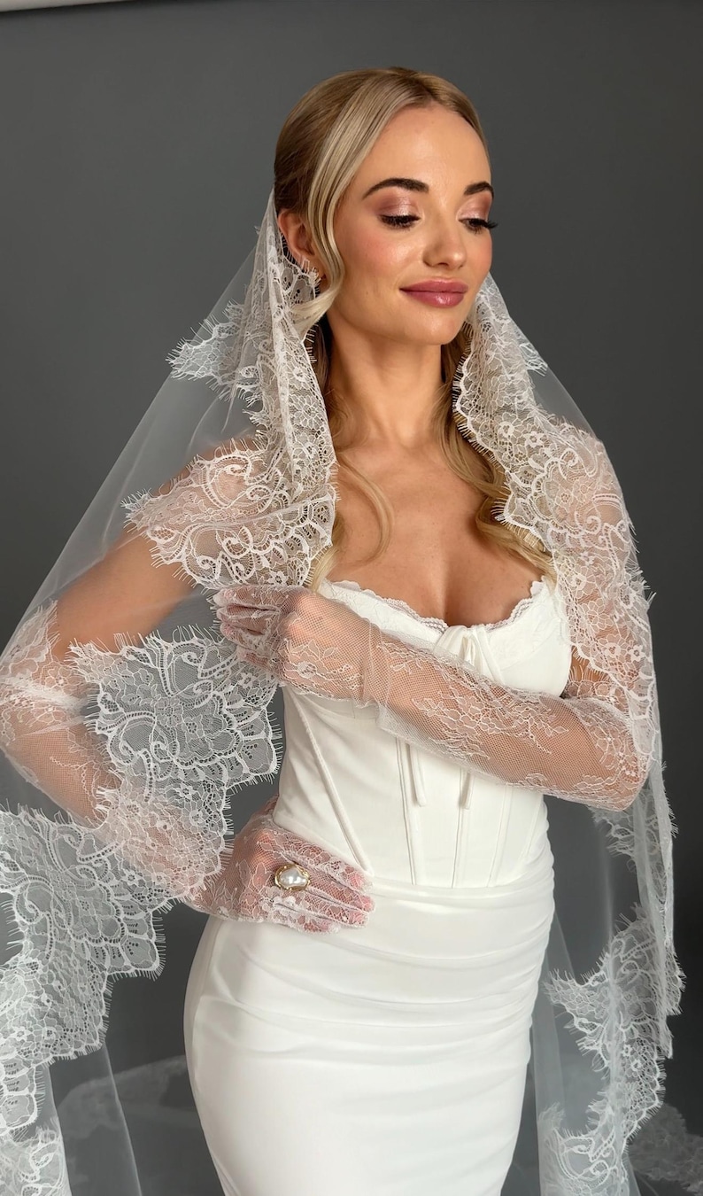 May include: A white wedding dress with a corset bodice and a fitted skirt. The bride is wearing a lace veil and lace gloves. The gloves have a pearl ring on the ring finger. The veil and gloves have a delicate floral lace design.