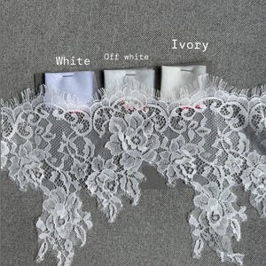 May include: A wide strip of white floral lace trim with scalloped edges, displayed alongside three small fabric swatches labelled White, Off white, and Ivory. The lace features an intricate botanical pattern with delicate mesh detailing.
